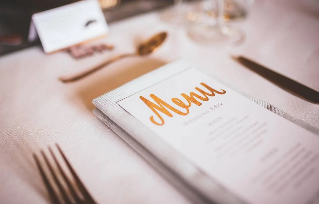 A copper-foil 'Menu' card on a wedding place setting at Vaulty Manor, with a wedding BBQ menu inside, place card, gold cutlery and a linen napkin on a pale tablecloth.
