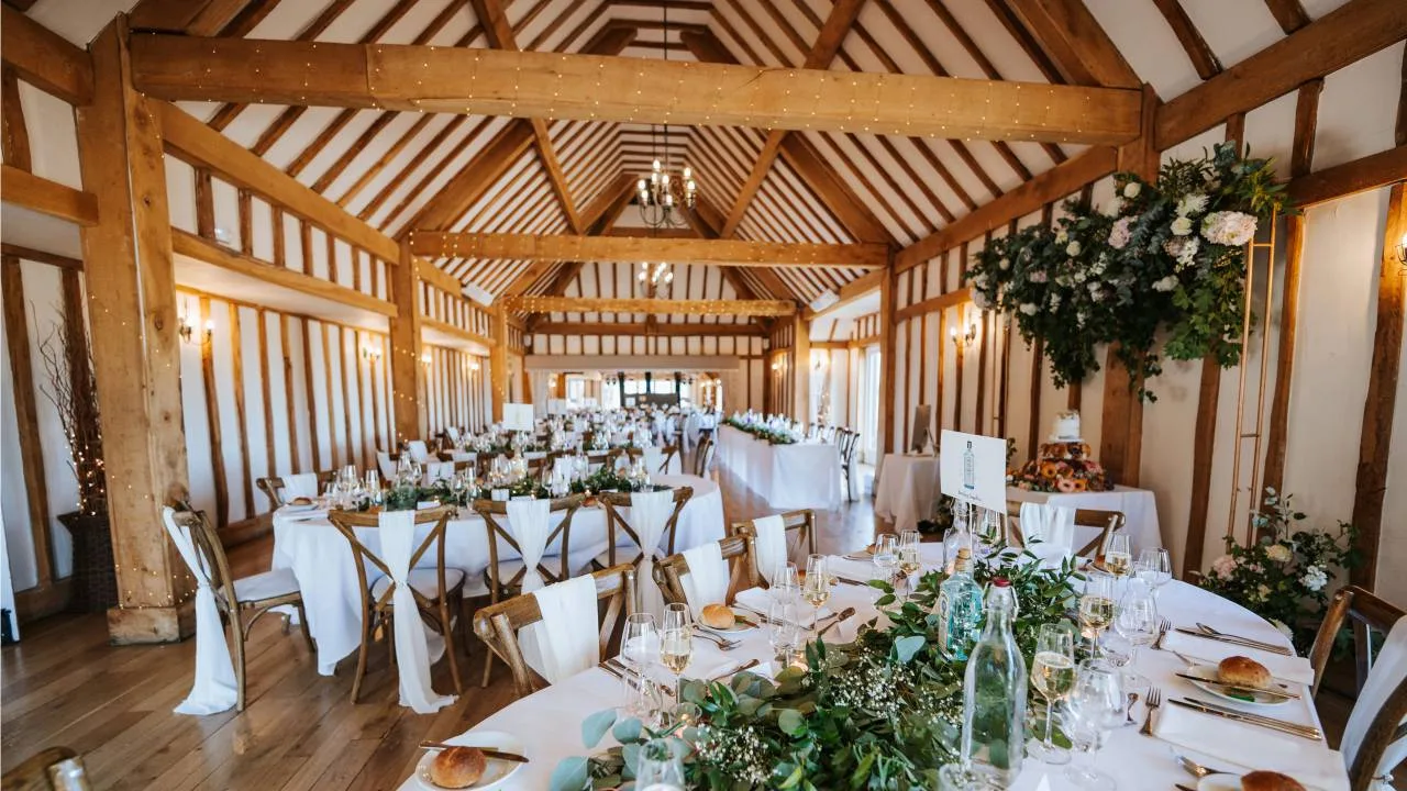 Inside the restored 17th-century barn at Vaulty Manor, dressed for a wedding breakfast with round tables, white linens, eucalyptus garlands, fairy-lit oak beams and a vaulted timber-framed roof above.