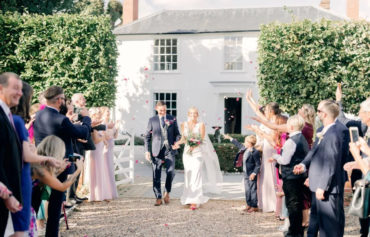 A bride and groom walking down a confetti-lined gravel aisle in front of the Manor House at Vaulty Manor, guests on both sides throwing petals on a sunlit afternoon.