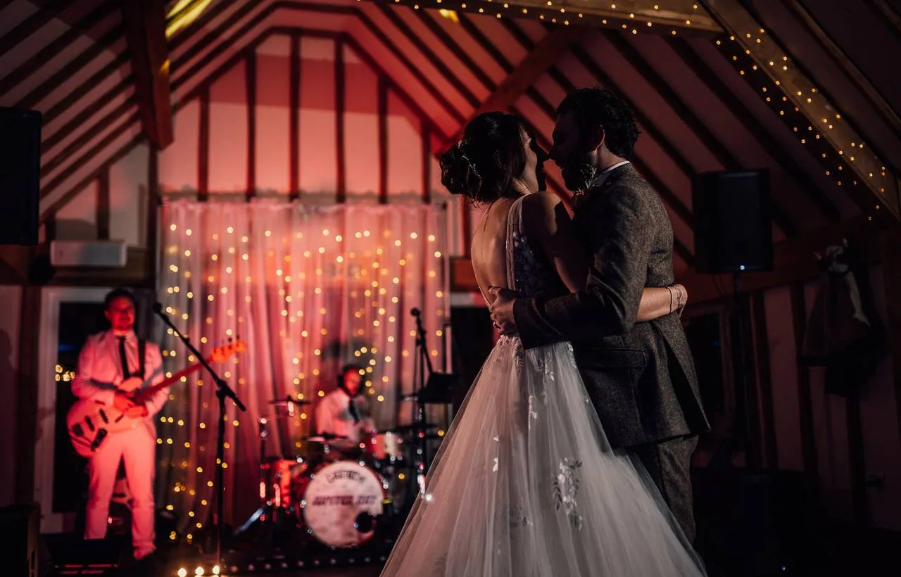 A bride and groom sharing their first dance in the candlelit barn at Vaulty Manor, a live band playing behind them under fairy-lit oak beams.
