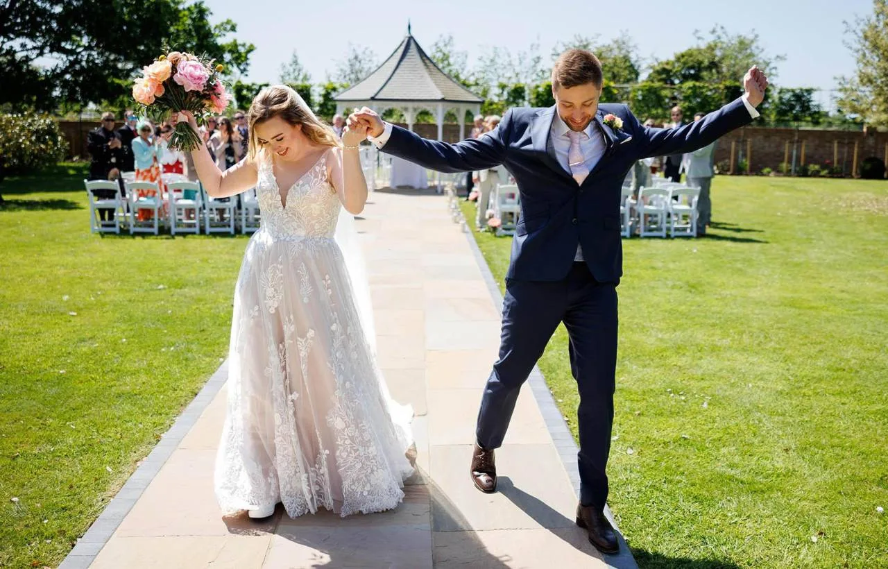 A bride and groom walking back down the aisle in the walled garden at Vaulty Manor, with the white pergola and seated guests behind them on a sunlit summer afternoon.