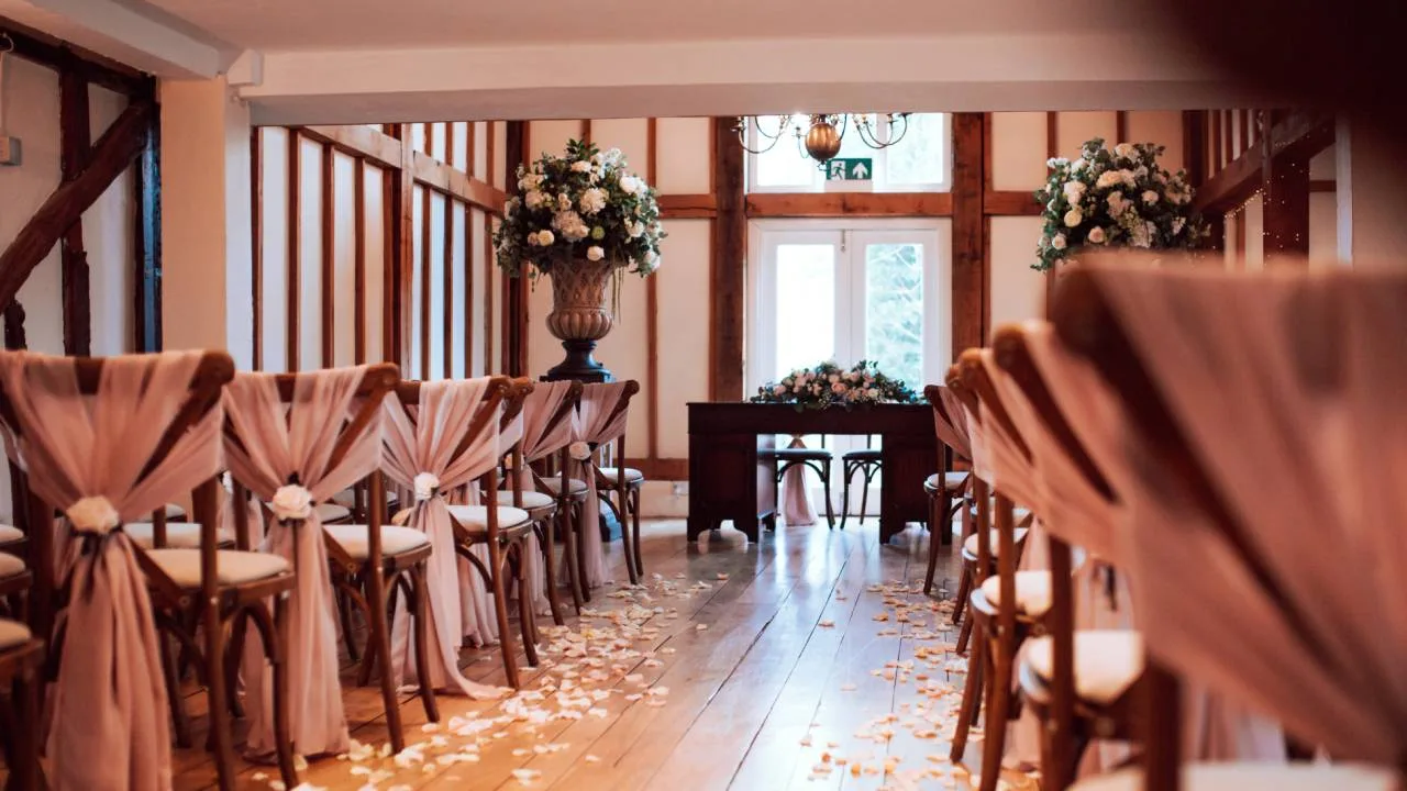 The barn at Vaulty Manor set up for a wedding ceremony, with cross-back chairs in dusty-pink sashes, a rose-petal-lined aisle and tall floral urns either side of the registrar's table.