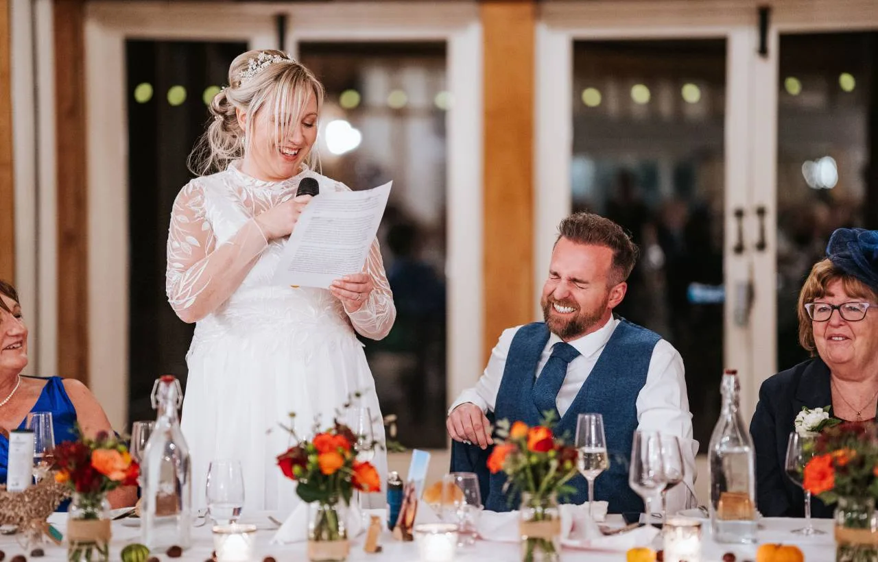 A bride giving a speech with a microphone during the wedding breakfast in the candlelit barn at Vaulty Manor, the groom laughing beside her at a long table of guests with autumn floral centrepieces.